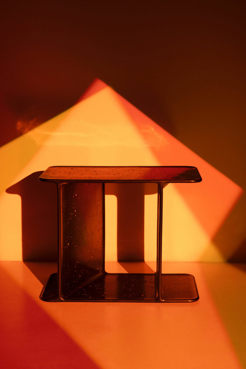 Una pequeña mesa rectangular de metal Holly Hunt se encuentra debajo de un patrón de luz cálido y geométrico que proyecta sombras nítidas en las paredes y el piso.