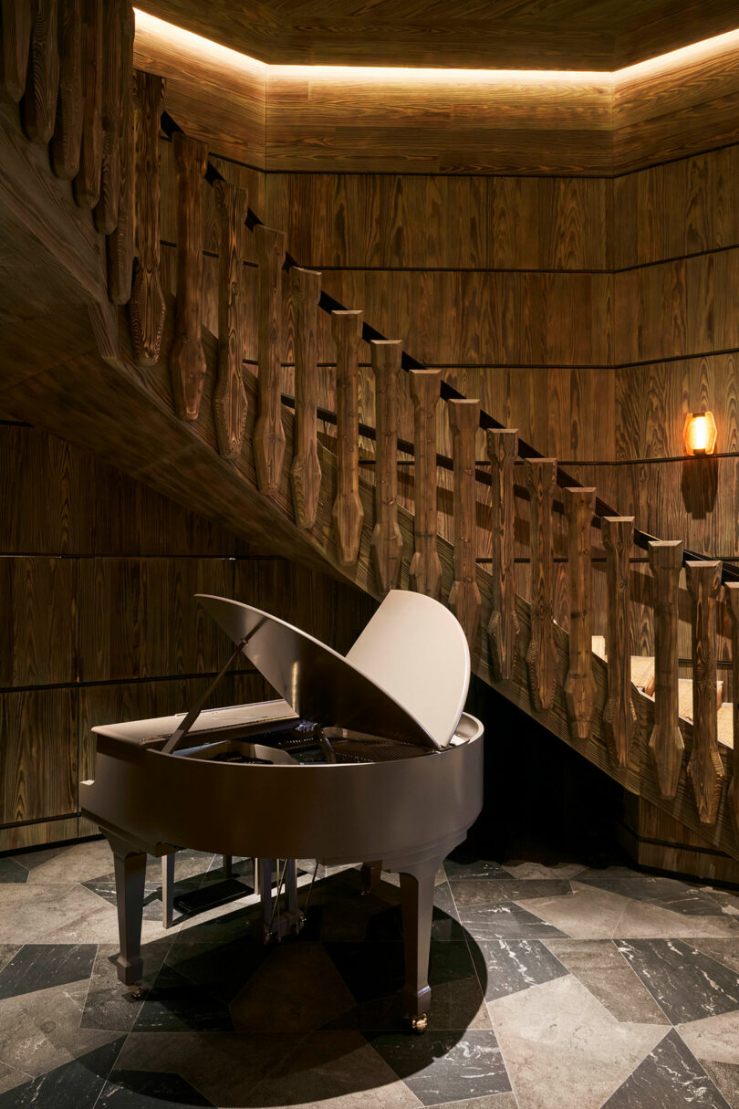 A white grand piano sits on a patterned stone floor near a wooden spiral staircase with carved railings in a dimly lit room.