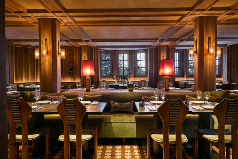 A warmly lit restaurant interior with wooden beams, green upholstered seating, set tables, red lamps, and large windows in the background.