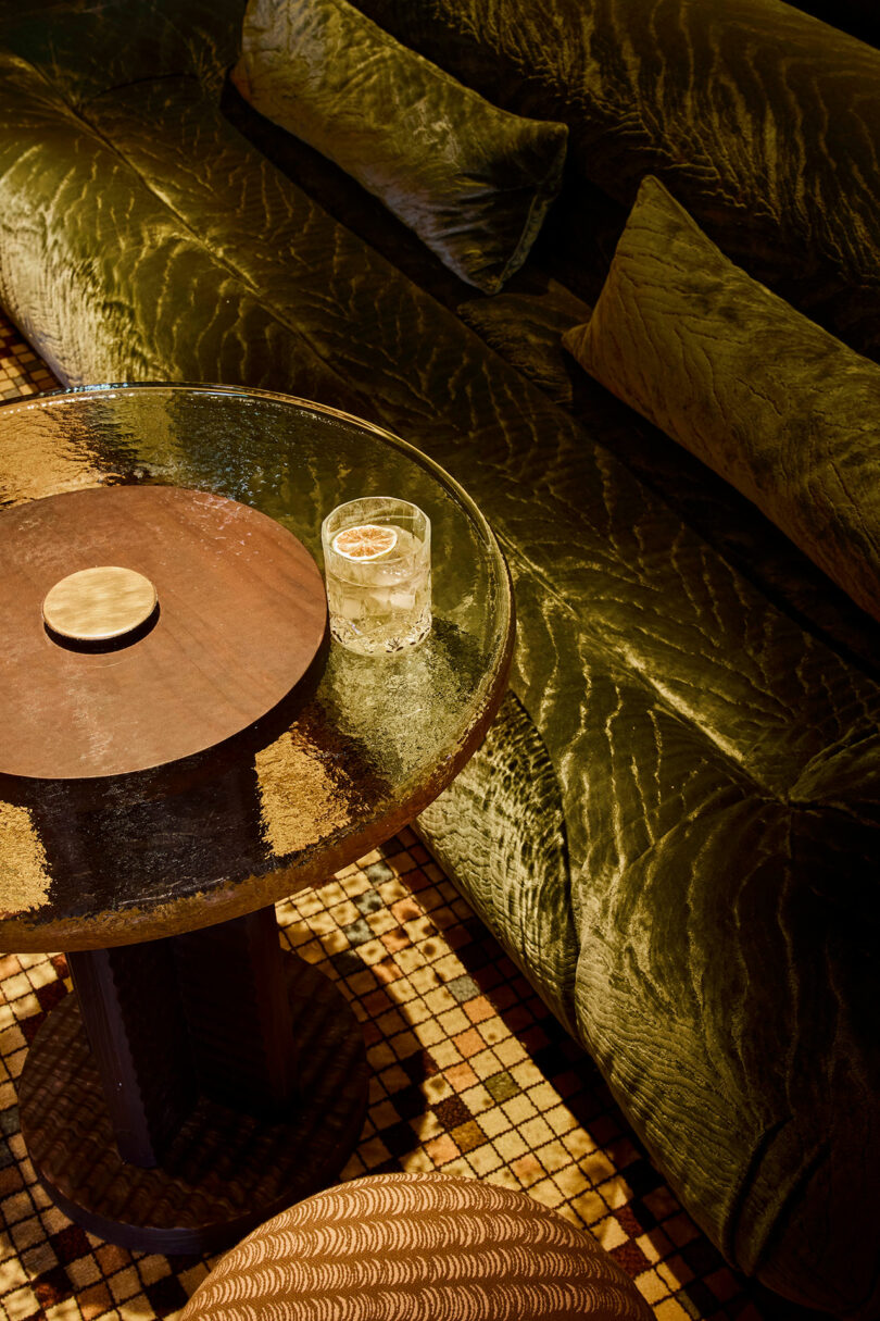 A glass of clear beverage with ice and a citrus slice sits on a round glass-topped table next to a green velvet sofa and a patterned stool on a tiled floor.