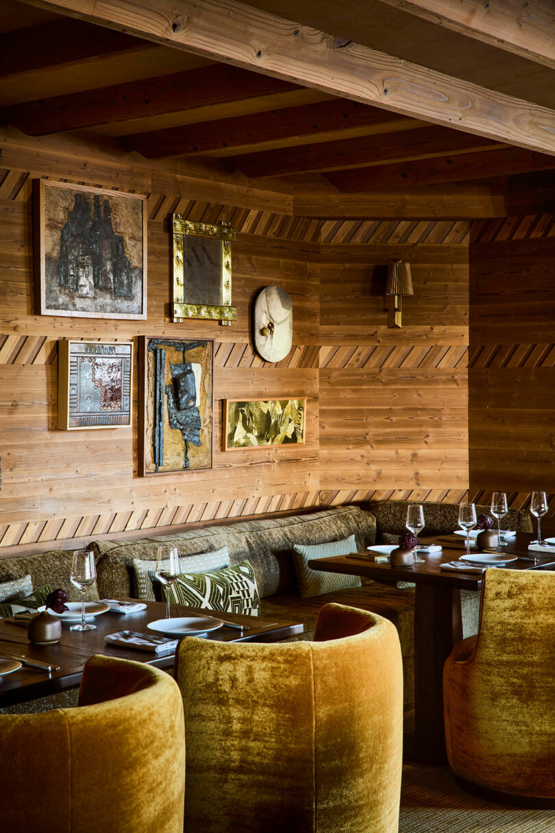A cozy restaurant interior with wood-paneled walls, mustard yellow velvet chairs, cushioned benches, set tables, and framed artwork on the walls.