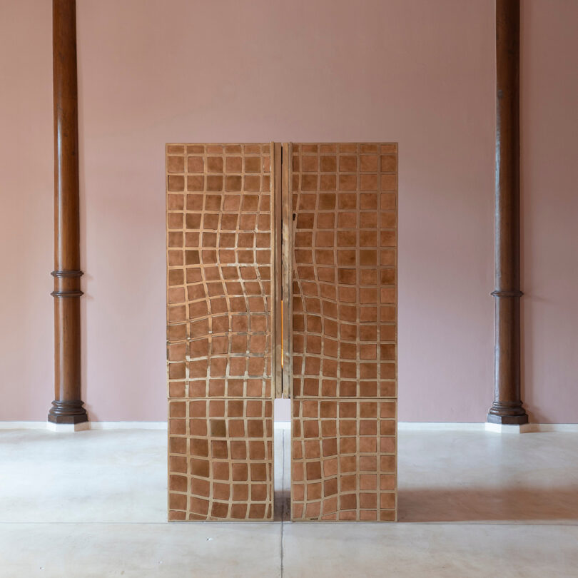 A wooden cabinet with a wavy, mesh pattern—a nod to Kelly Wearstler's signature style—stands in the center of a room with two wooden columns and a pink wall in the background.