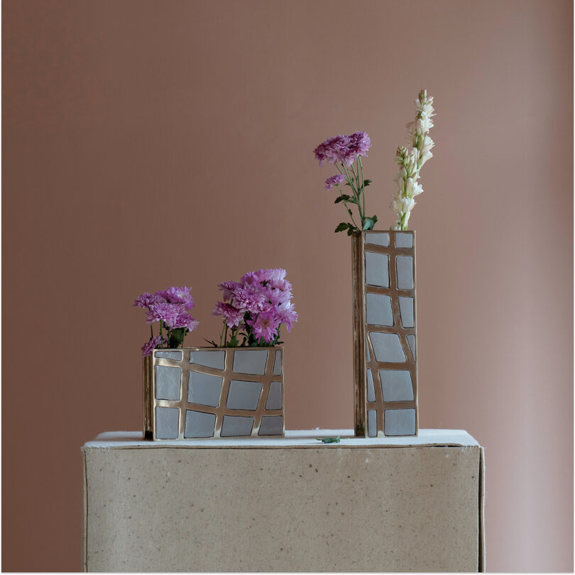 Three geometric vases with silver and gold accents, reminiscent of the style of Kelly Wearstler, hold purple and white flowers, placed on a light pedestal against a brown background.