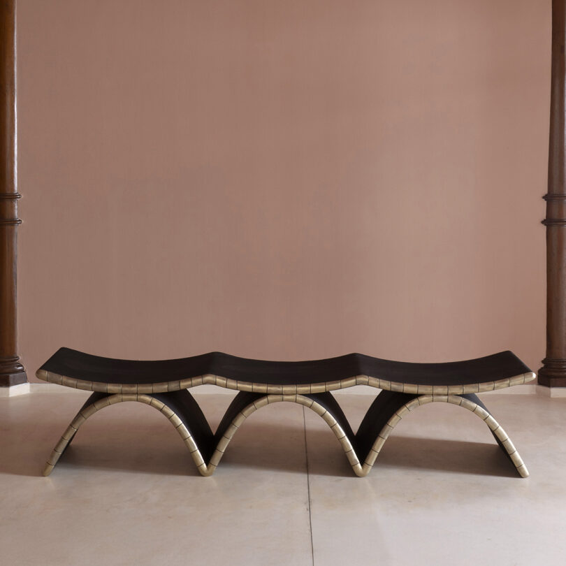 A sculptural bench with an undulating, wavy form and dark surface, supported by light, curved legs, channels Kelly Wearstler's signature style on a simple purple wall and light floor.