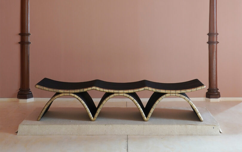 A Kelly Wearstler-inspired sculptural bench with an undulating black seat and gold-edged arches sits on a stone platform in a minimalist room with pink walls and two wooden columns.