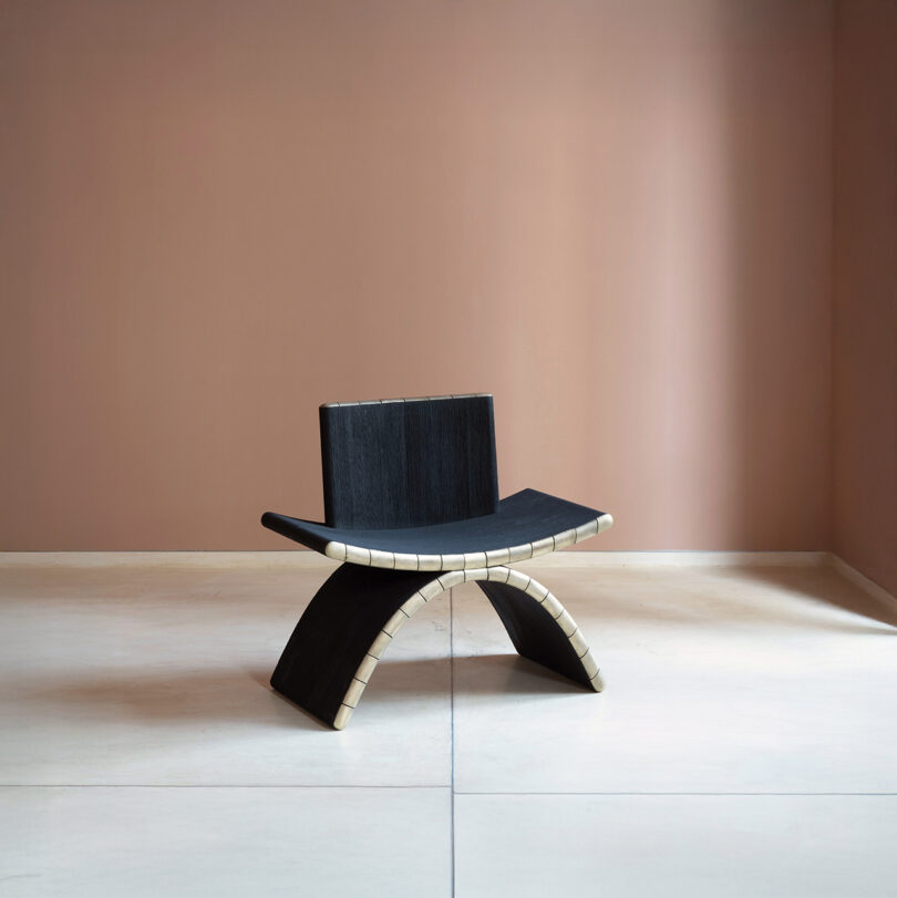 A modern black chair with a curved arch-like base, inspired by Kelly Wearstler, sits on a light floor, with beige accents against a muted pink wall.