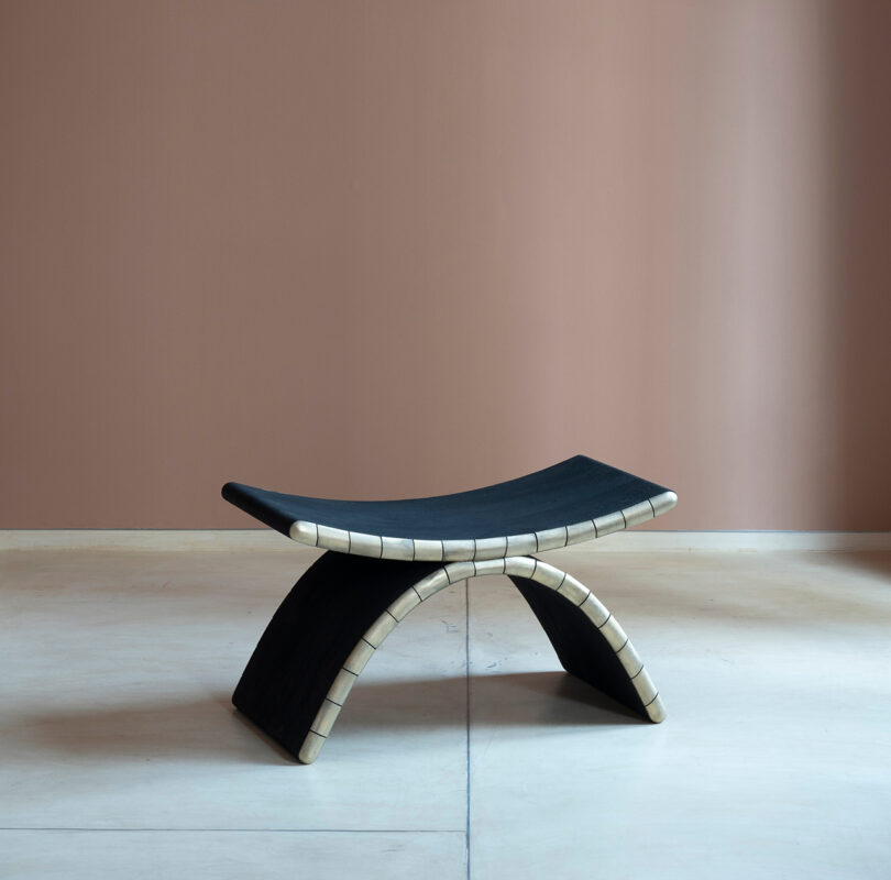 A modern Kelly Wearstler-inspired black bench with a curved seat and arched base with light accents, set against a rose-brown wall and light floor.