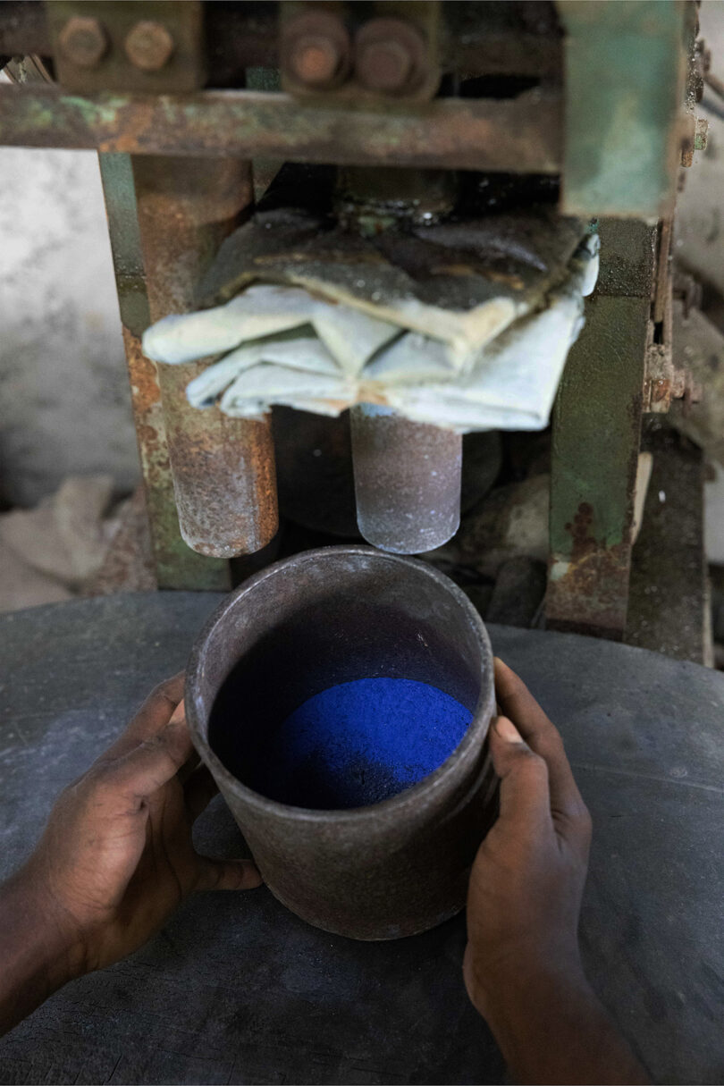 A person holds a metal container filled with blue powder under a pressing machine with folded cloths on top, evoking Kelly Wearstler's bold, tactile aesthetic.