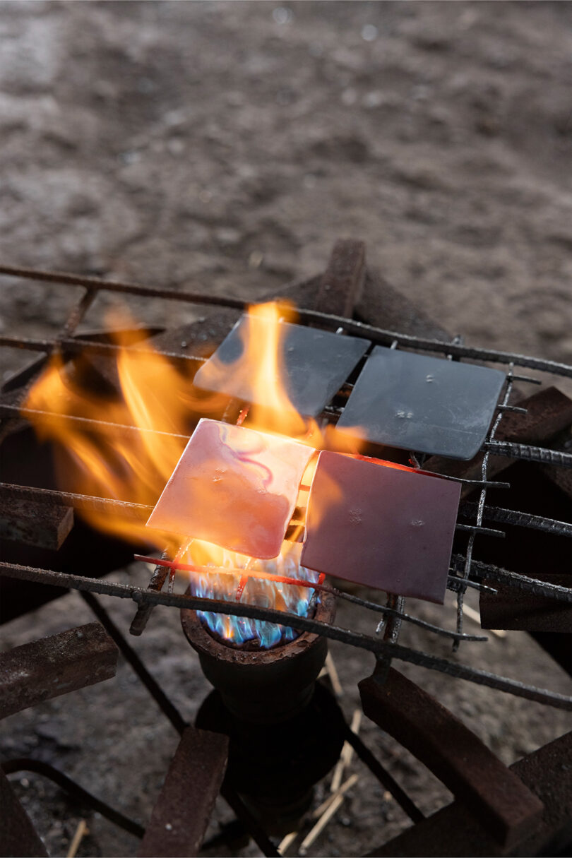 Four metal squares heat over a gas burner with visible blue and orange flames, placed on an outdoor metal grill - evoking a bold, sculptural look reminiscent of Kelly Wearstler's iconic design style.