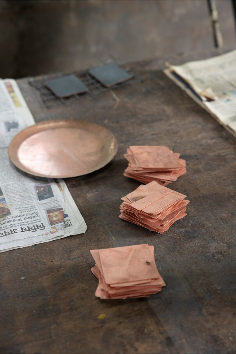 Stacks of copper sheets and a round copper plate, reminiscent of Kelly Wearstler's bold style, are placed on a wooden table with some newspapers nearby.