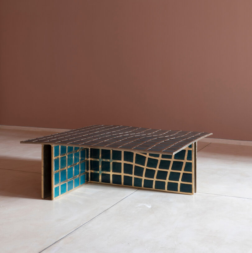 A modern Kelly Wearstler-inspired coffee table with an uneven grid pattern in dark brown and gold, with blue tiles on the sides, on a minimalist pink wall and light floor.