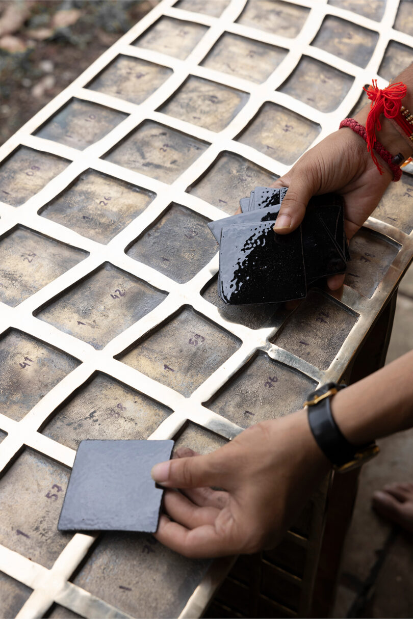 Two hands place glossy black tiles on a metal grid of numbered squares, reminiscent of Kelly Wearstler's bold, modern style.