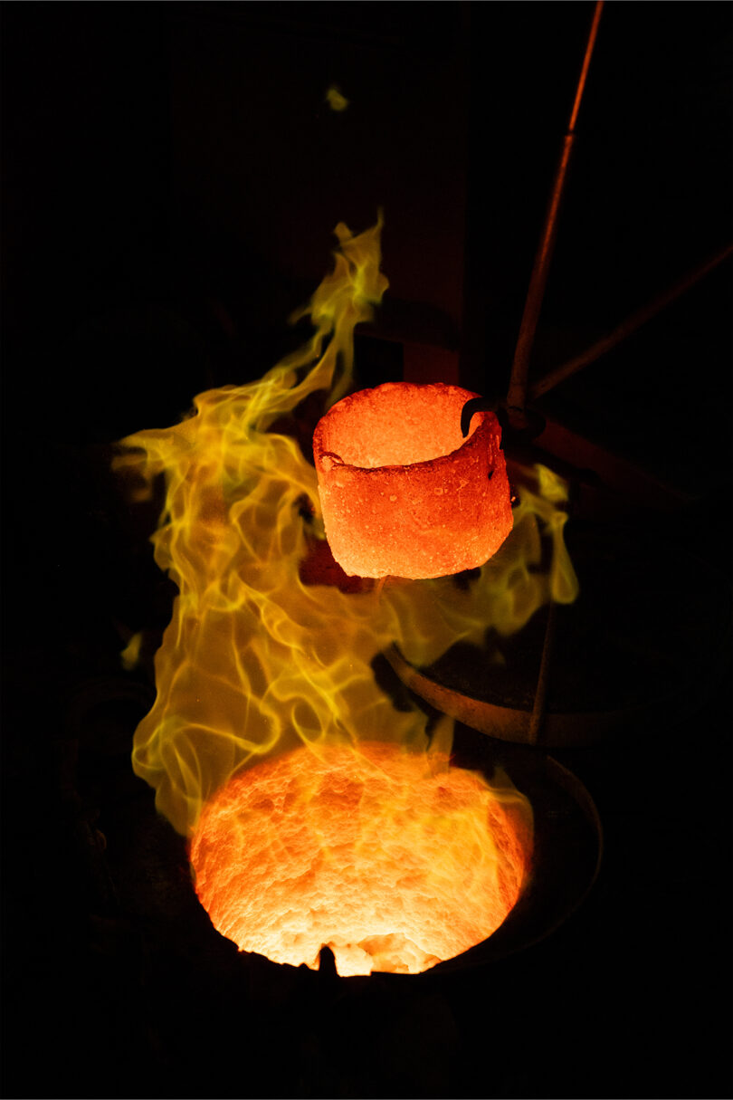 A glowing crucible filled with molten metal rises above a furnace, with yellow flames rising around it—a scene that echoes Kelly Wearstler's bold, dramatic style.