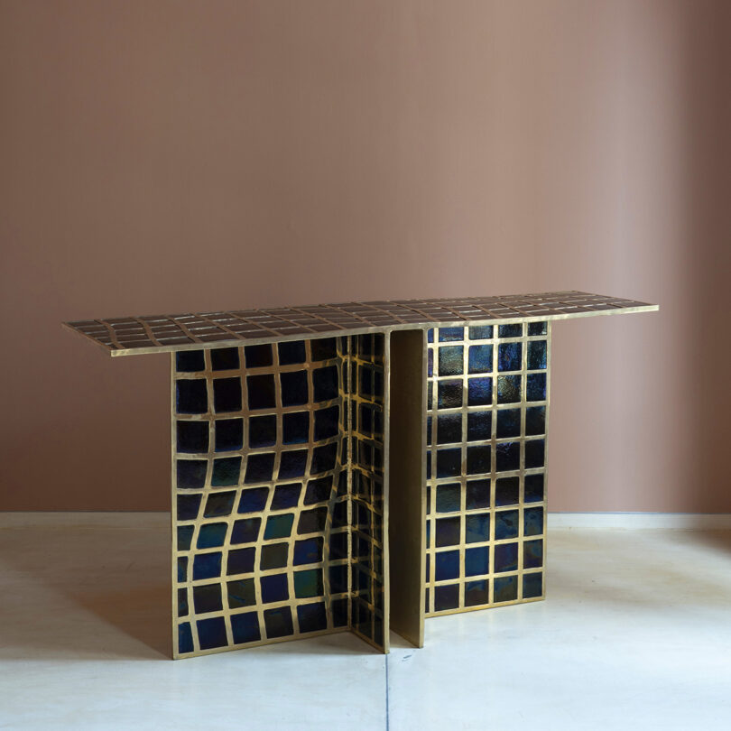 A rectangular console table with a gold metal frame and a wavy grid pattern of dark blue and gold tiles, reminiscent of Kelly Wearstler's signature style, on a simple beige wall and open floor.