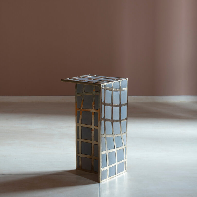 A small, modern Kelly Wearstler-inspired side table with a geometric grid pattern in gray and gold stands on a plain, pale floor against a muted purple wall.