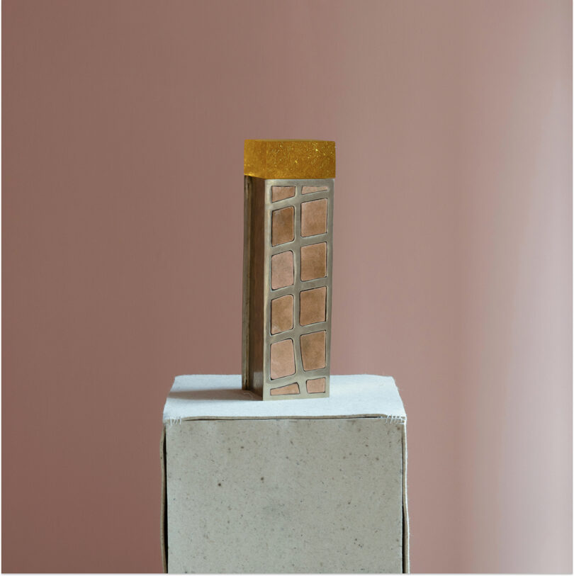 A rectangular object with a gold top and geometric metallic sides, reminiscent of Kelly Wearstler's signature style, sits on a textured white pedestal against a muted pink background.