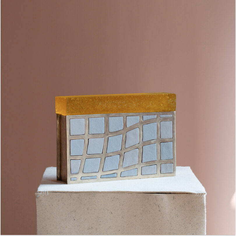 A rectangular object with a gold top and a silver, irregular grid pattern on the front, reminiscent of Kelly Wearstler's signature style, rests on a textured beige platform on a pale pink background.