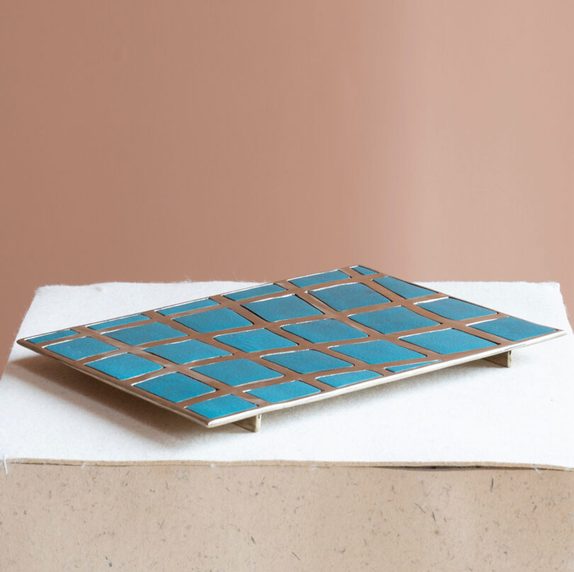 A rectangular tray with a grid of blue tiles and brass edges, inspired by the bold style of Kelly Wearstler, displayed in a white surface on a beige background.