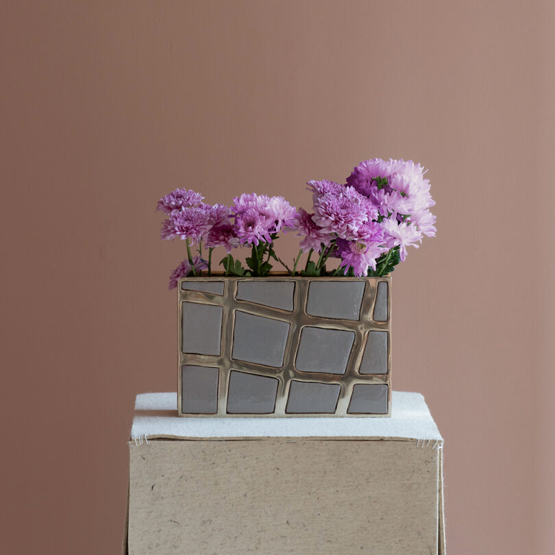 Kelly Wearstler-inspired rectangular vase with a geometric design holding light and dark purple flowers, placed on a fabric plinth against a plain purple background.