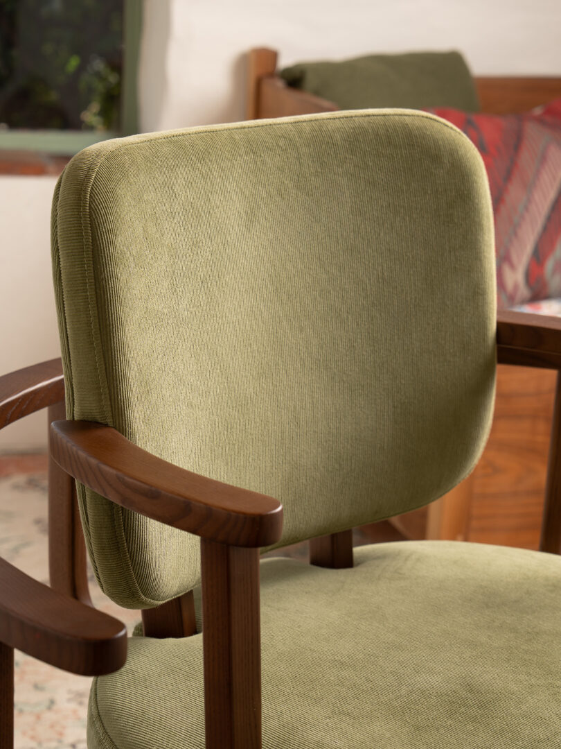Green upholstered chair with wooden arms and legs placed in a room with a bed and nightstand in the background.