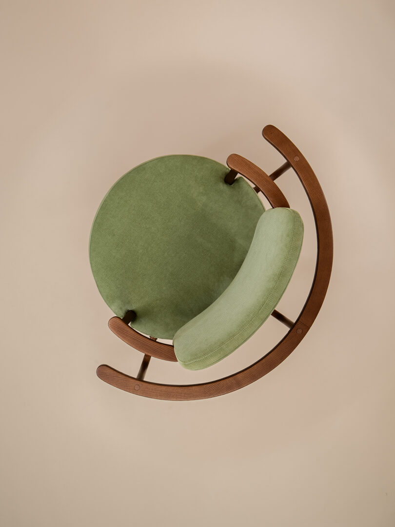 Top view of a modern chair with a circular green seat and curved wooden frame on a beige background.