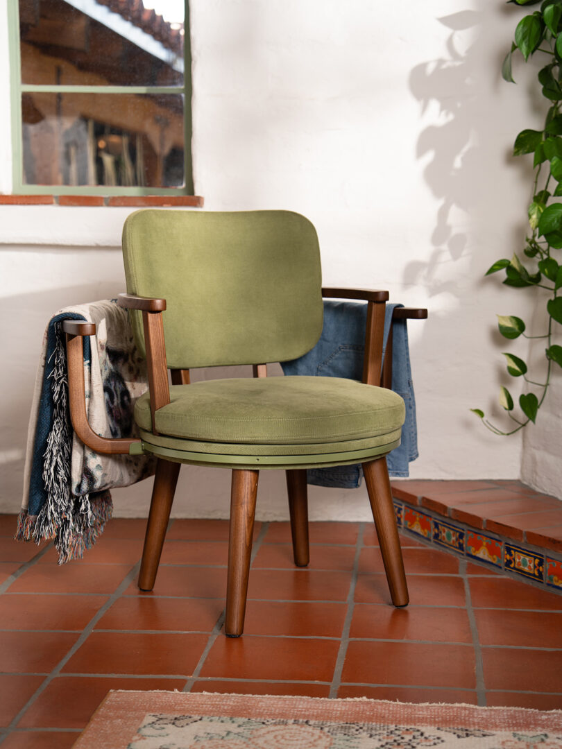 A green wooden chair with cushions sits on a tiled floor near a wall, with blankets draped over its arms and a leafy plant hanging nearby.