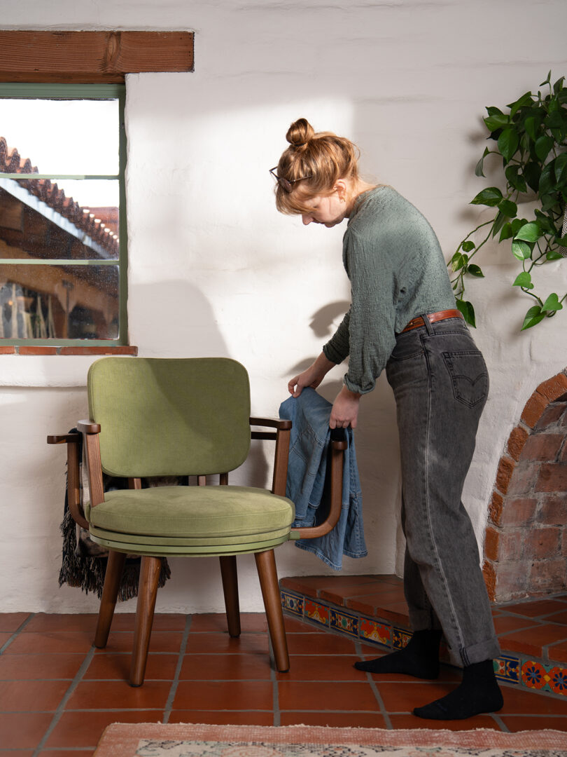 A woman stands next to a green chair in a sunny room, putting on a denim jacket on the arm of the chair.
