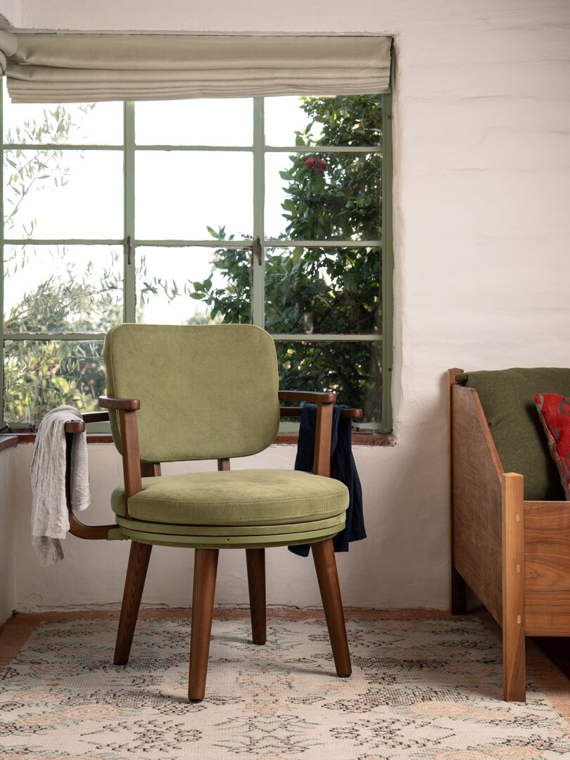 A green wooden chair with cushions sits on a patterned rug near a window with greenery outside. a white scarf and blue garment hang from the arms of the chair.