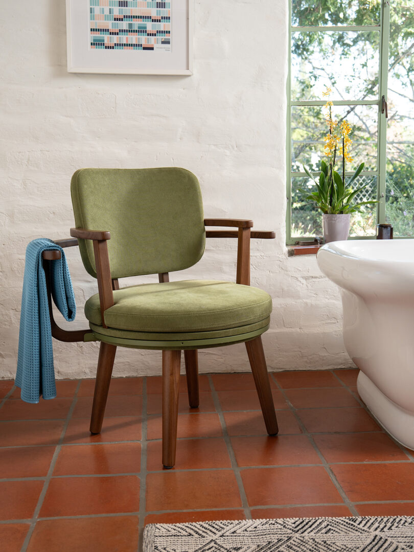 A green wooden chair with cushions is placed on a terracotta tiled floor next to a bathtub. a blue towel hangs over the arm of the chair. A window and a flower pot are visible in the background.