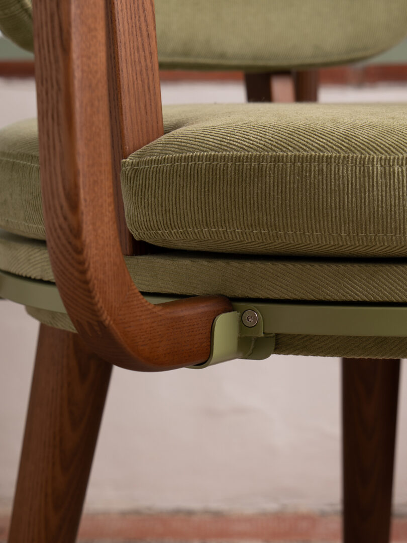 Close up of chair with green corduroy cushion, wooden legs and armrest and green metal support bracket under the seat.
