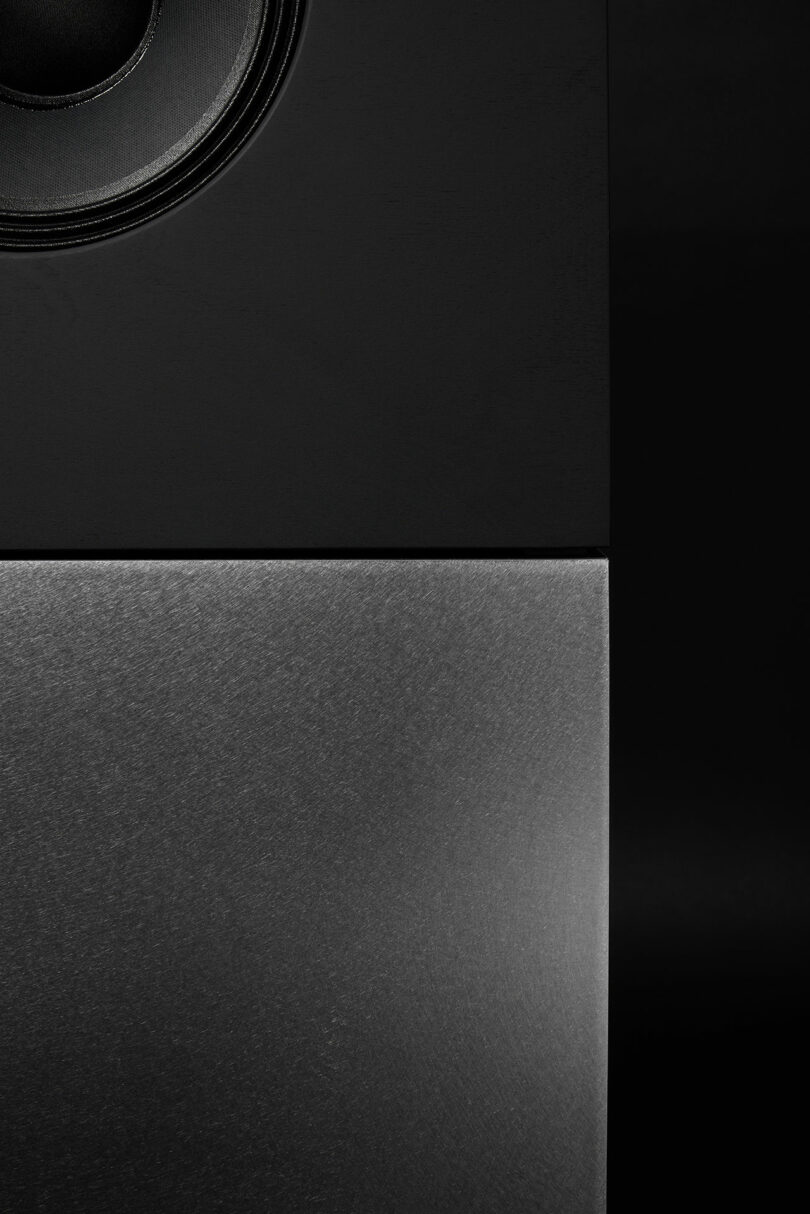 Close-up of the Braque stereo system, a collaboration by Studio D.A and Nocs, showing a black speaker with a visible round driver at the top left and a brushed metal panel below, photographed in low light.