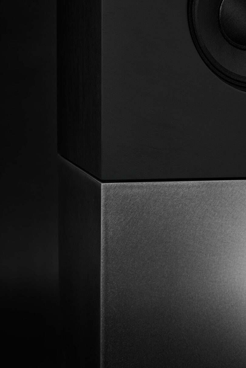 Close-up of a modern black speaker from Studio D.A and Nocs' debut Braque stereo system, featuring a matte upper section, a brushed metal lower section, and part of the speaker driver visible on the top right.