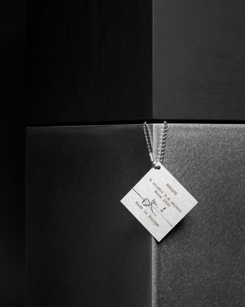 A square metal tag with text and a ball chain hangs against a background of dark, brushed metallic surfaces—echoing the refined design language seen as Studio D.A and Nocs debut the Braque stereo system.