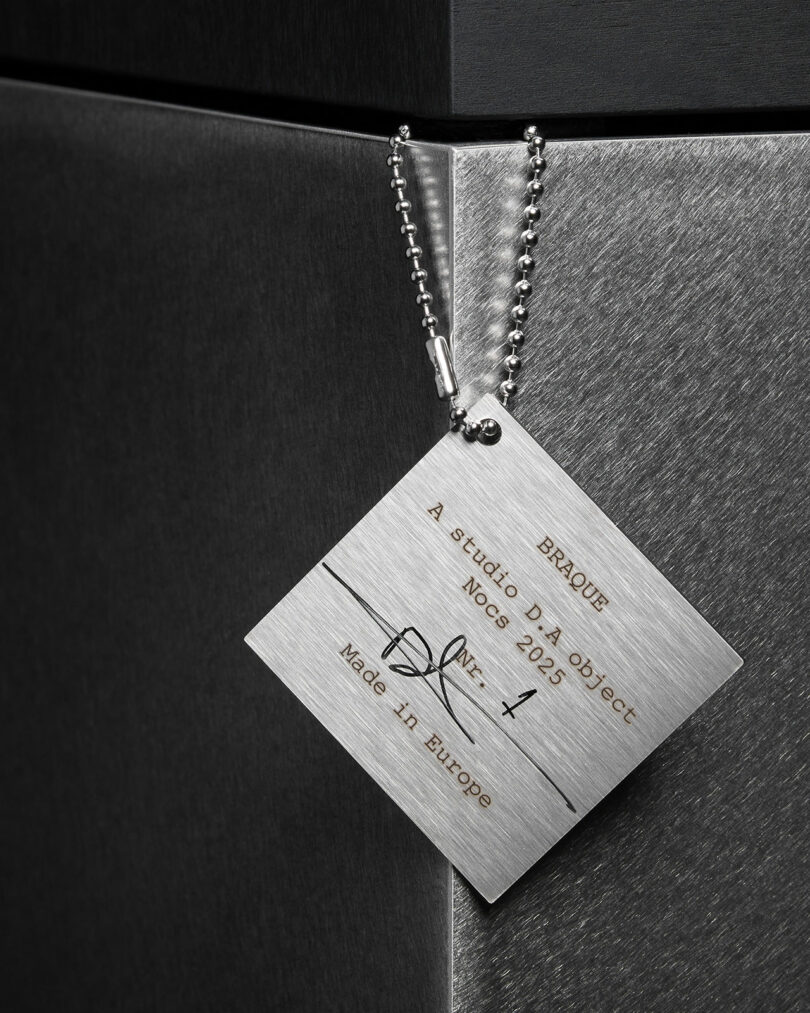 A metal tag with a ball chain is attached to a metallic surface, displaying text about the object, including "BRAQUE," "No: 1," "2025," and "Made in Europe”—marking Studio D.A and Nocs' debut of the Braque stereo system.