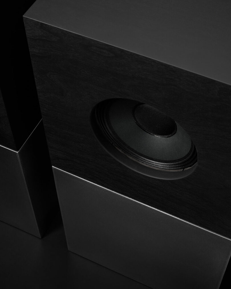 Close-up view of a modern black speaker with a visible woofer, featuring sharp lines and geometric shapes against a dark background, as Studio D.A and Nocs debut the Braque stereo system.