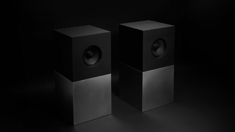 Studio D.A and Nocs debut the Braque stereo system: two minimalist, cube-shaped speakers with black upper halves containing the drivers and metallic lower halves, set against a dark background.