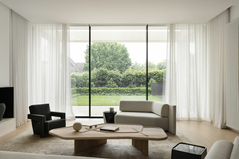 Minimalist living room with light wood floors, modern furniture, a large wooden coffee table, and floor-to-ceiling windows overlooking a green garden.