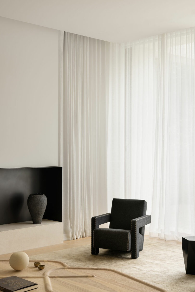 Minimalist living room with a black armchair, light rug, a black vase on a low shelf, and floor-to-ceiling sheer white curtains letting in natural light.