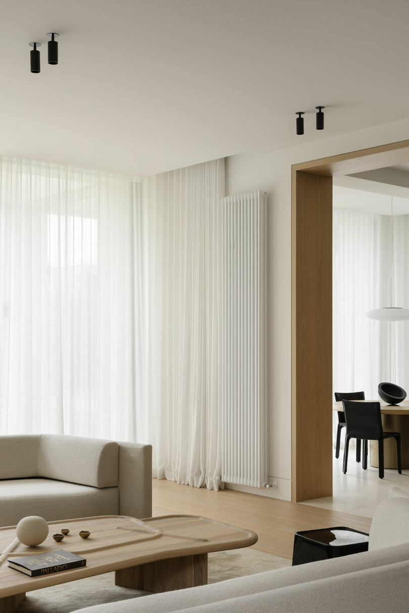 Minimalist living room with light-colored sofas, wooden coffee table, floor-to-ceiling sheer curtains, and a view into a dining area with black chairs and a round white pendant lamp.