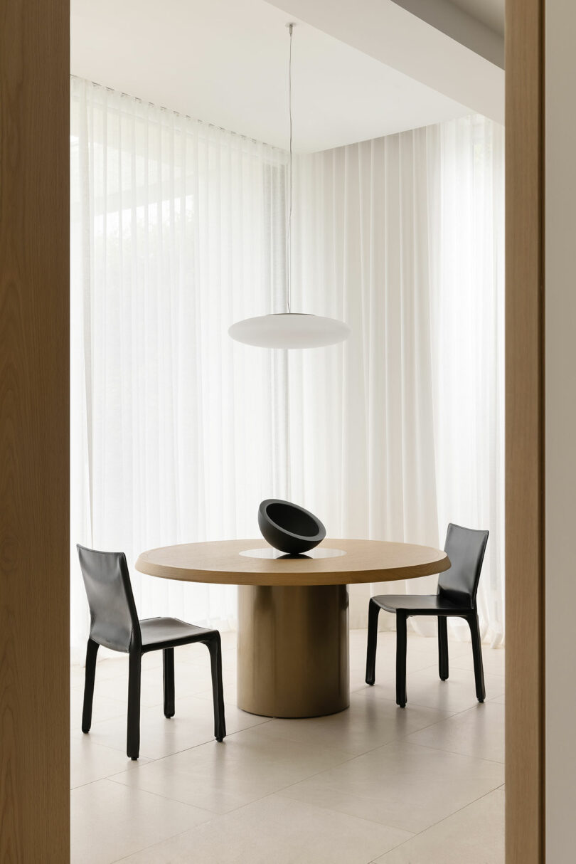 A round wooden table with a black sculptural centerpiece is flanked by two black chairs in a minimalist room with sheer white curtains and a suspended white light fixture.