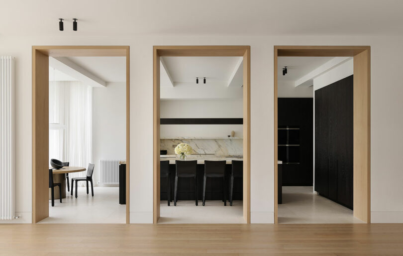Minimalist interior featuring three large wooden-framed doorways leading to a dining area, kitchen with marble backsplash, and a dark cabinetry storage area.