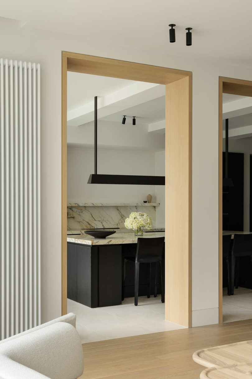 Modern kitchen seen through a light wood doorway, featuring a marble island, black chairs, pendant light fixture, and a vase of white flowers on the counter.
