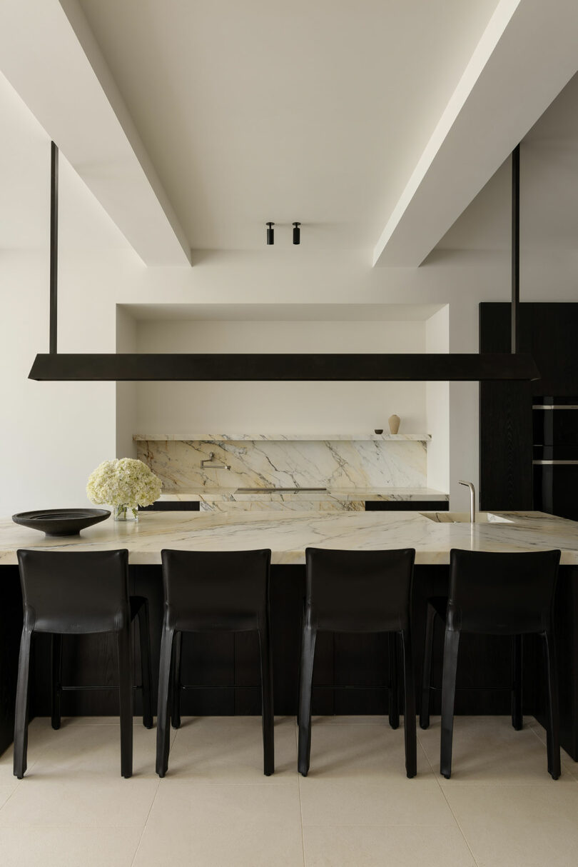 Modern kitchen with a marble island, four black chairs, a long black pendant light, and a marble backsplash. A bowl of white flowers sits on the counter.