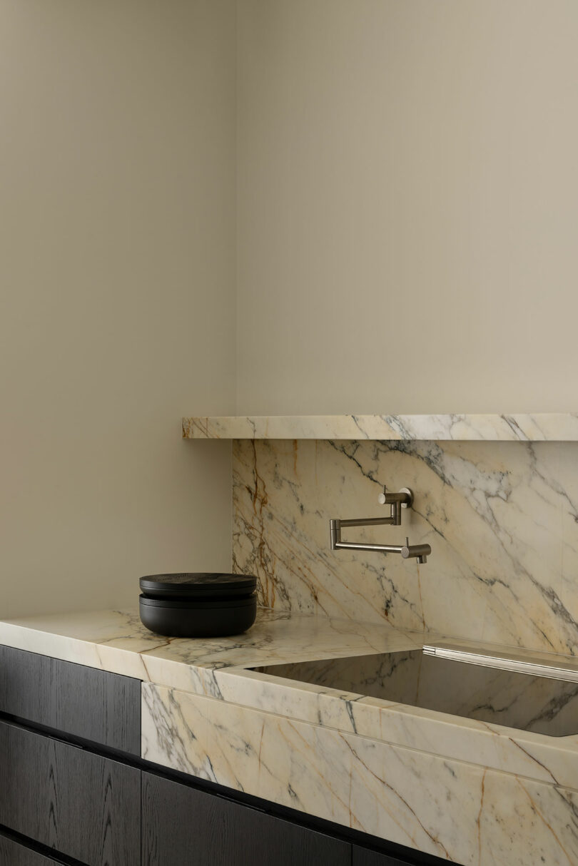 A minimalist bathroom features a marble countertop and backsplash, a wall-mounted faucet, a black round vessel, and dark wood cabinetry.