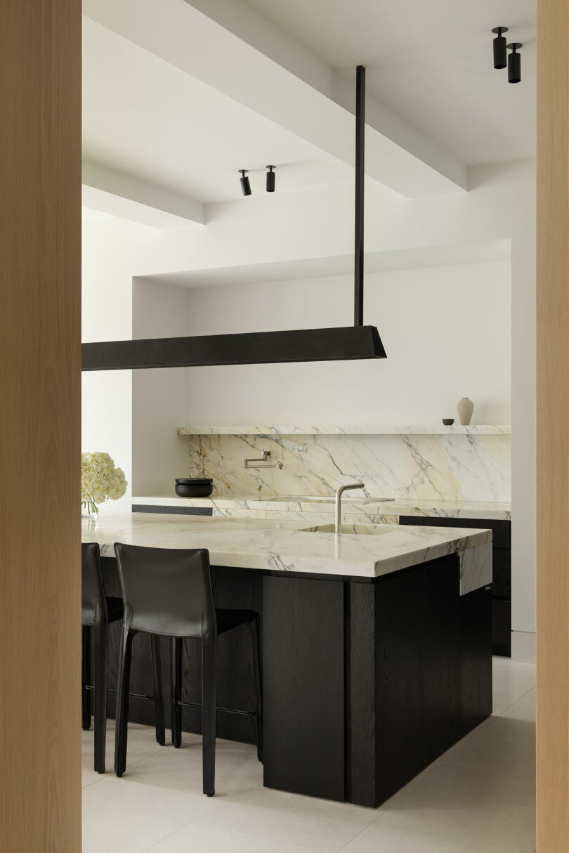 Modern kitchen with black cabinetry, a marble island and backsplash, minimalist black chairs, and neutral decor, viewed through a doorway.