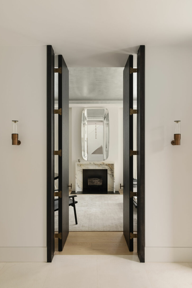 Modern living room with double black doors, marble fireplace, large wall mirror, and minimalist decor in neutral tones.