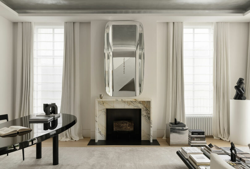 Modern living room with a marble fireplace, large abstract mirror above, black sculptural decor, books, and a glossy black table with an open book, all set against white walls and tall windows.