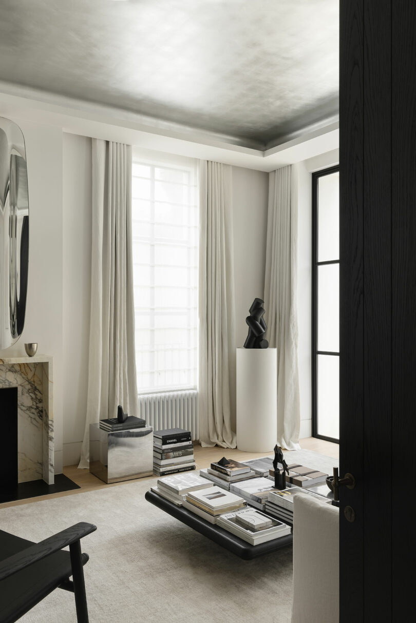 Minimalist living room with neutral tones, a low coffee table stacked with books, a marble fireplace, sheer curtains, and a modern black sculpture on a pedestal near a large window.