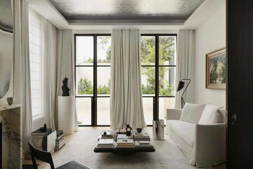 Minimalist living room with floor-to-ceiling windows, white sofa, marble fireplace, coffee table with books, modern art sculpture, and neutral decor.