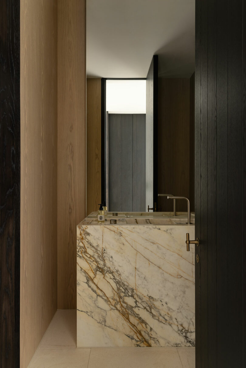 Minimalist bathroom with light wood walls, a marble bathtub with brown and gold veining, a modern faucet, and a partially open dark wood door.